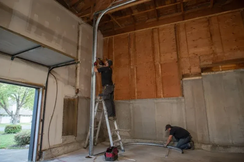 One person on a stepladder working beside a tall curved garage-door track while another kneels beside loose track sections in an unfinished garage. — High-Lift Garage Door Conversion, Kitchener