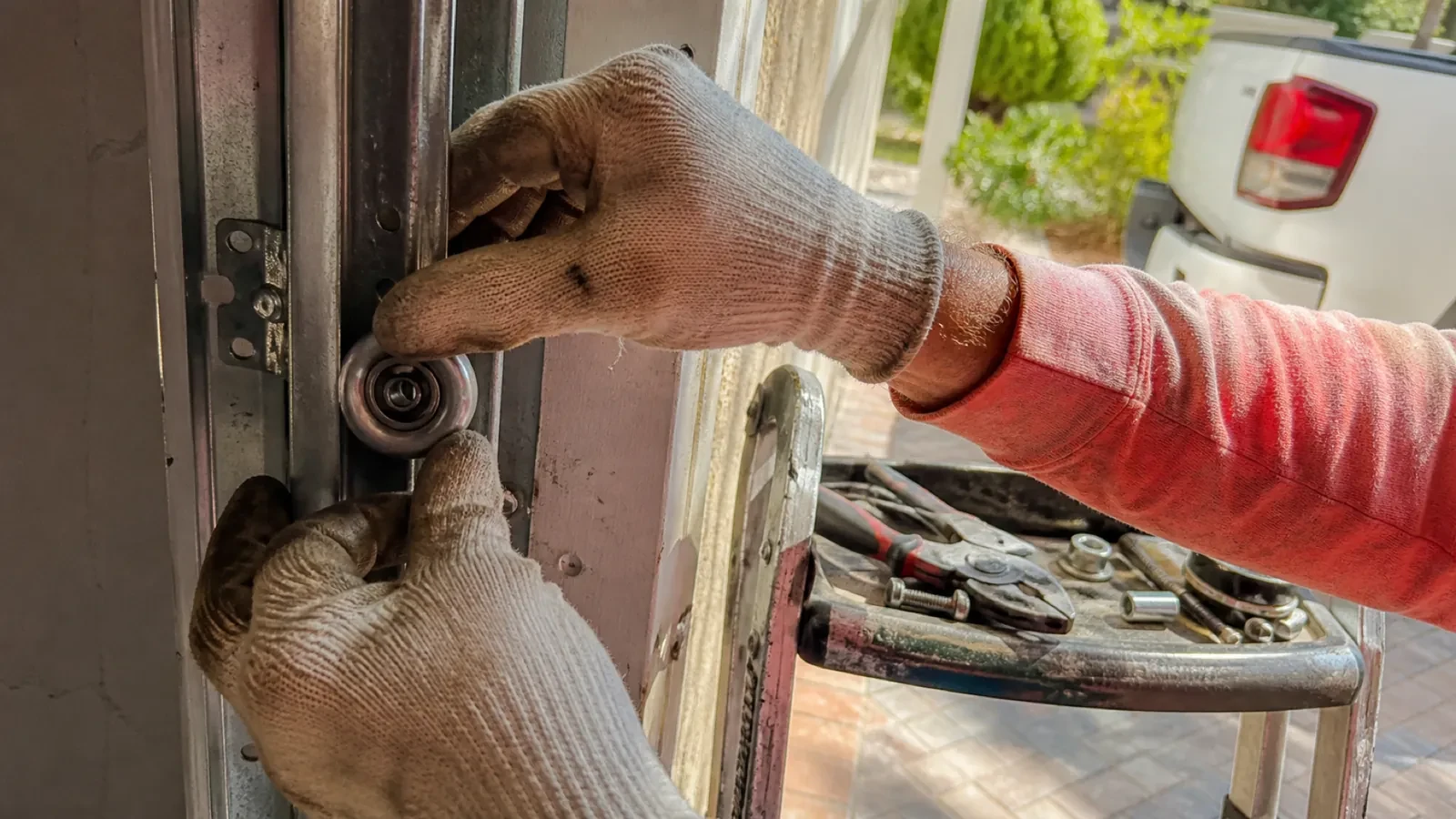Gloved hands holding a metal roller wheel against a door track, with pliers and small hardware on a stepladder tray nearby. — Country Hills