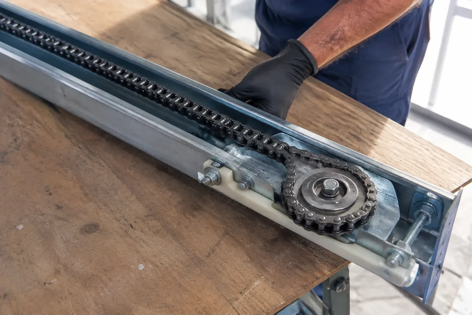 Gloved hand resting on a steel opener rail laid on a wooden bench, showing a drive chain looped around a sprocket wheel. — Chain-Drive Opener Repair, Kitchener